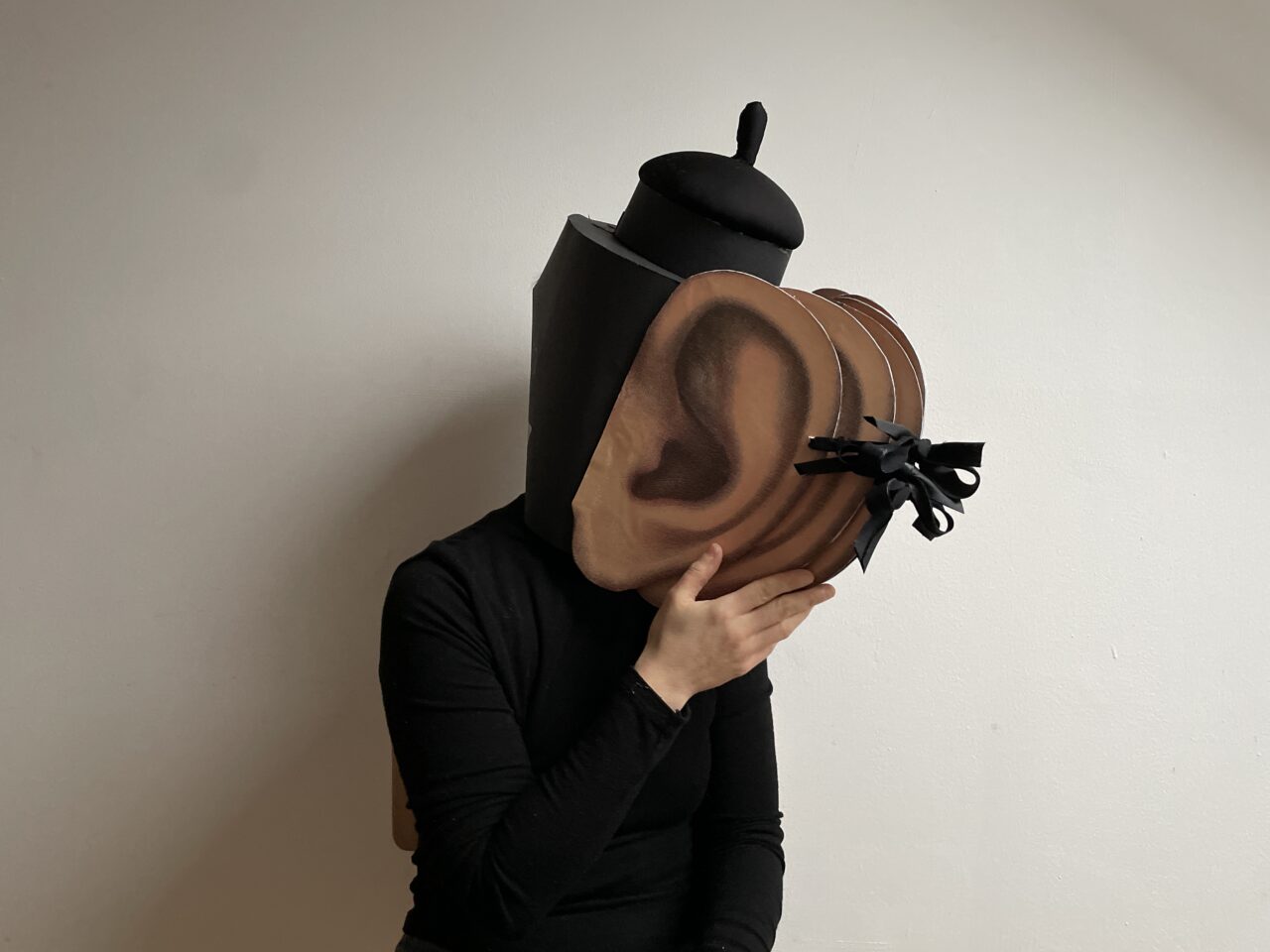Eine Person trägt ein surrealistisches Kopfkostüm mit überdimensionalen braunen Ohren und schwarzer Kopfbedeckung.