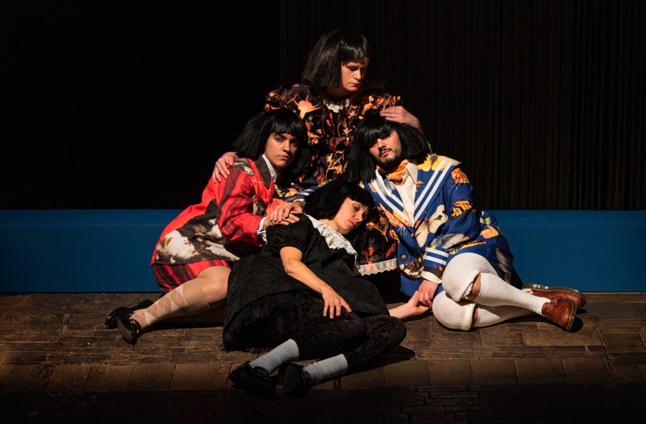 Auf einer Theaterbühne sitzen vier Personen eng beieinander auf dem Boden. Sie tragen auffällige, bunte Kostüme und kurze schwarze Perücken. Eine Person liegt vorne und wirkt erschöpft oder schläft. Die drei anderen sitzen dicht um sie herum. Sie halten und stützen die liegende Person vorsichtig. Die Gruppe wirkt ruhig, nachdenklich und verbunden. Das Licht ist warm und richtet sich auf die Personen. Der Hintergrund ist dunkel, die Bühne aus Holz.