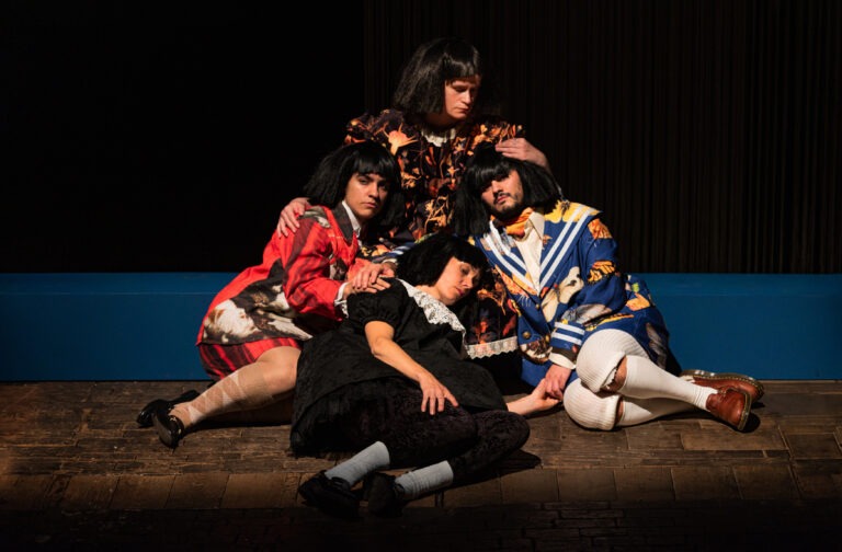 Auf einer Theaterbühne sitzen vier Personen eng beieinander auf dem Boden. Sie tragen auffällige, bunte Kostüme und kurze schwarze Perücken. Eine Person liegt vorne und wirkt erschöpft oder schläft. Die drei anderen sitzen dicht um sie herum. Sie halten und stützen die liegende Person vorsichtig. Die Gruppe wirkt ruhig, nachdenklich und verbunden. Das Licht ist warm und richtet sich auf die Personen. Der Hintergrund ist dunkel, die Bühne aus Holz.