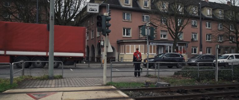 Das Bild zeigt eine städtische Straßenszene an einer Kreuzung mit Straßenbahngleisen im Vordergrund. Eine Person mit rotem Rucksack steht auf dem Gehweg und wartet offenbar an einer Ampel. Links fährt ein großer roter LKW vorbei, während auf der gegenüberliegenden Straßenseite mehrere Autos unterwegs oder geparkt sind. Im Hintergrund befinden sich mehrstöckige Wohnhäuser mit einer Fassade in warmen Farbtönen. Bäume ohne Blätter deuten auf eine kühle Jahreszeit hin. Ein Verkehrsschild mit der Aufschrift „Vorsicht!“ und einem Straßenbahnsymbol weist auf die nahegelegenen Gleise hin. Die Szene vermittelt einen alltäglichen Moment im urbanen Straßenverkehr.