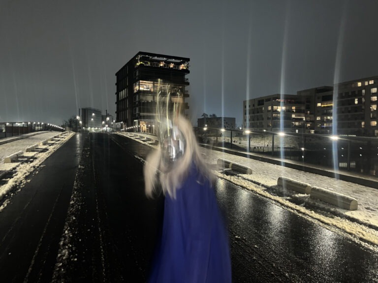 Nachts steht eine Person auf einer nassen, dunklen Straße, die von Schnee gesäumt ist. Sie trägt eine silberne Maske und ein langes blaues Kleid oder einen Mantel. Durch die Bewegung wirkt die Figur unscharf und leicht verschwommen. Im Hintergrund sind moderne, mehrstöckige Gebäude mit beleuchteten Fenstern zu sehen. Straßenlampen werfen lange Lichtstreifen in den Himmel und spiegeln sich auf dem glatten Asphalt. Die Szene wirkt kühl, urban und leicht geheimnisvoll.