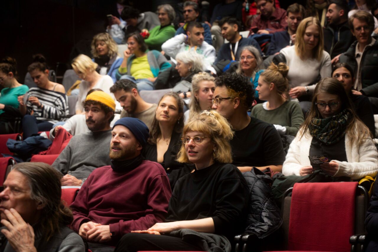 Publikum in einem Kinosaal oder Theater, dicht besetzte Reihen mit Menschen, die gespannt warten oder miteinander sprechen.